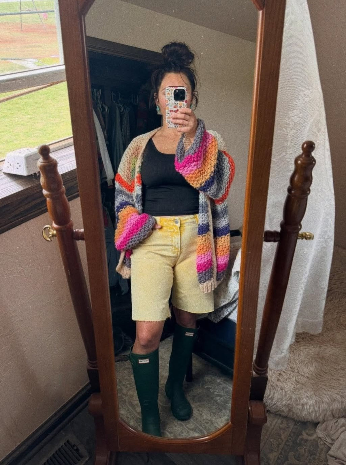 Person wearing a colorful cardigan, black top, yellow shorts, and green boots standing in front of a mirror.